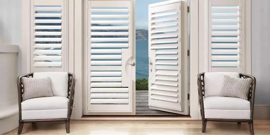 White shutters on French doors and side windows, one door open to an ocean view, between two white armchairs