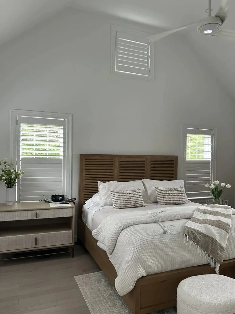 White plantation shutters on three bedroom windows, including a high gable window, above a wood bed and nightstand