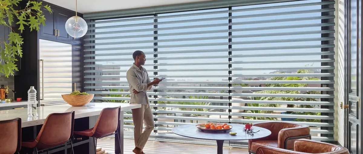 A man holding a phone stands by a wall of grey sheer horizontal shades in a modern kitchen with leather chairs