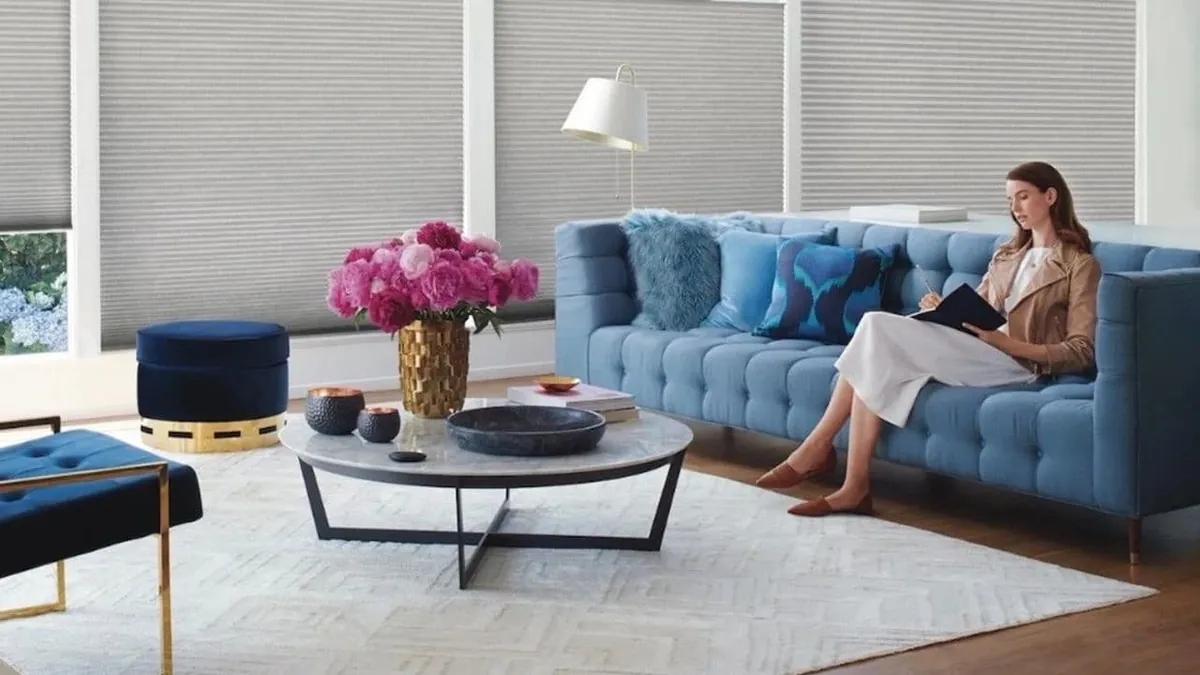 Grey honeycomb shades fully lowered behind a blue tufted sofa where a woman sits writing, with pink peonies on the coffee table