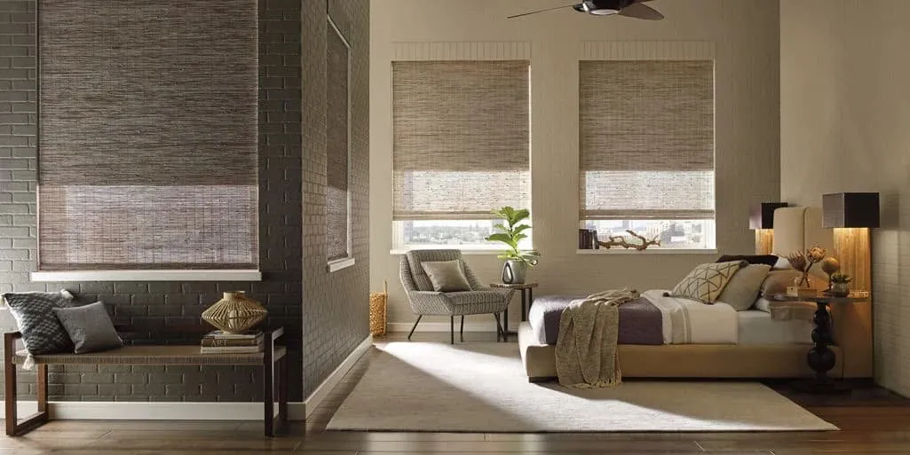 Woven wood shades in brown and grey tones on bedroom windows, with a bed, armchair and exposed brick wall