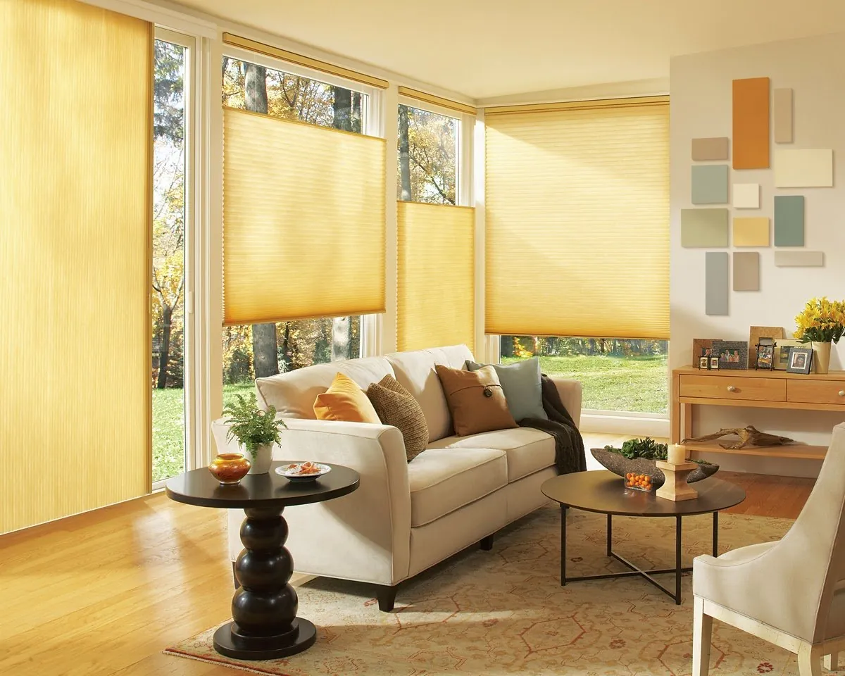 Golden honeycomb shades lowered from the top on corner windows and a vertical honeycomb shade on a sliding door, in a sunny living room with a cream sofa