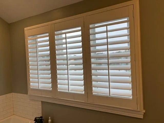 White plantation shutters with open louvres on a wide three-panel bathroom window above a white tiled tub