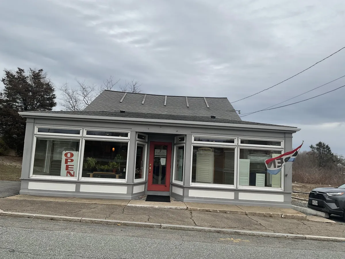 The Blinds and Designs Sagamore showroom from the street: a grey shop with large windows, a red front door, an Open sign and Hunter Douglas shutter samples in the window