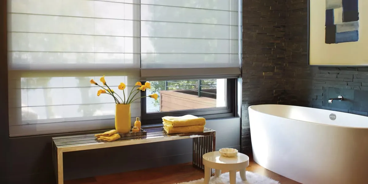 White sheer roman shades partly raised on windows in a bathroom with a freestanding tub, dark stone wall and yellow towels