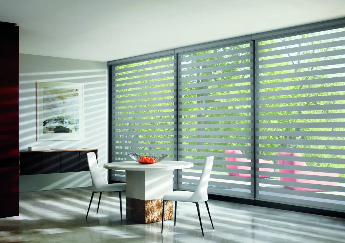 Grey banded shades with alternating sheer and solid stripes across a wall of tall windows, casting striped light over a white dining table and chairs