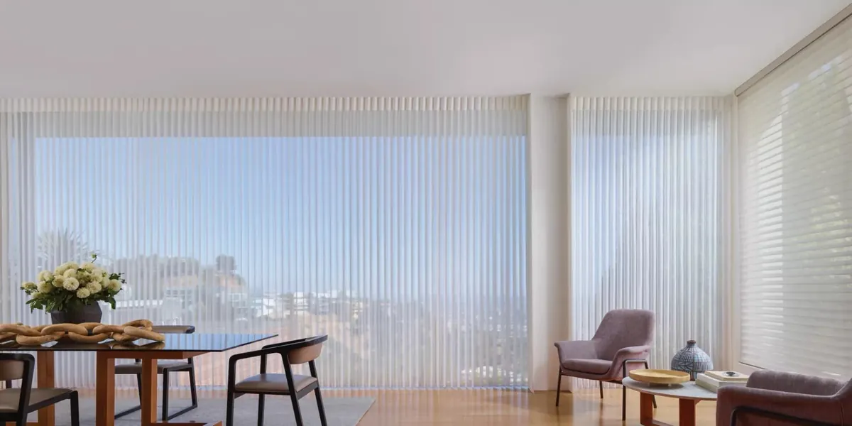 Floor-to-ceiling white vertical sheers across a wall of windows in an open dining and sitting room, with horizontal sheer shades on the side window