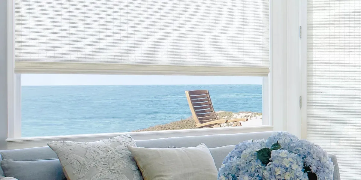 White woven shade raised partway on a window overlooking the sea and a wooden deck chair, behind a light blue sofa