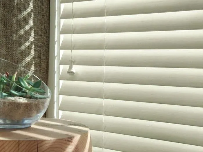 Close-up of white horizontal blind slats with a wood-grain texture and a cord tassel, beside a glass succulent bowl