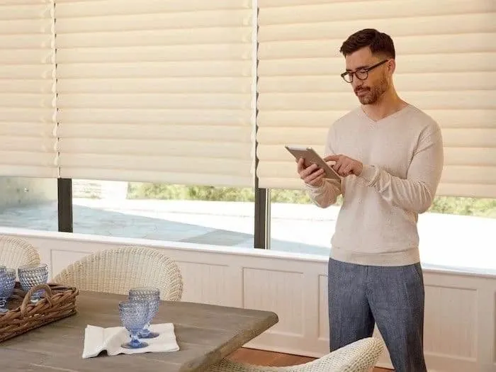 A man adjusting cream soft-fold shades from a tablet in a bright dining room with a wooden table and wicker chairs