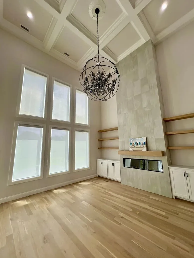 White roller shades lowered on six tall windows in a two-storey living room with a chandelier and tiled fireplace
