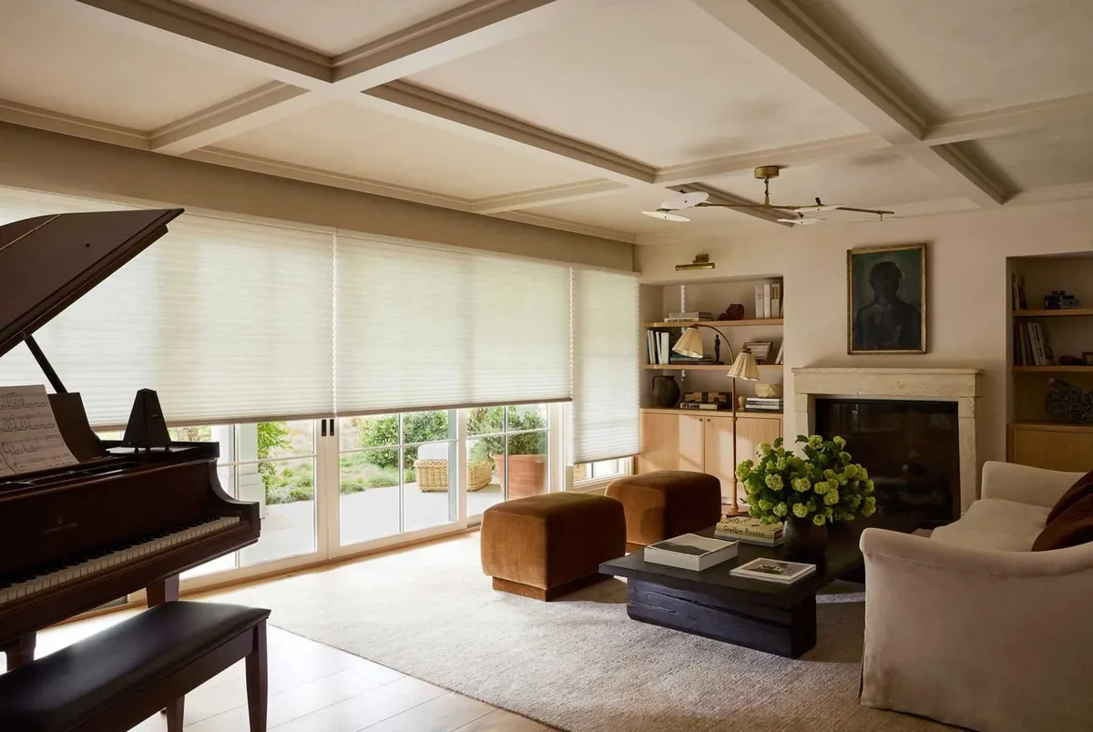Ivory honeycomb shades lowered across wide patio doors in a living room with a grand piano, coffered ceiling and stone fireplace