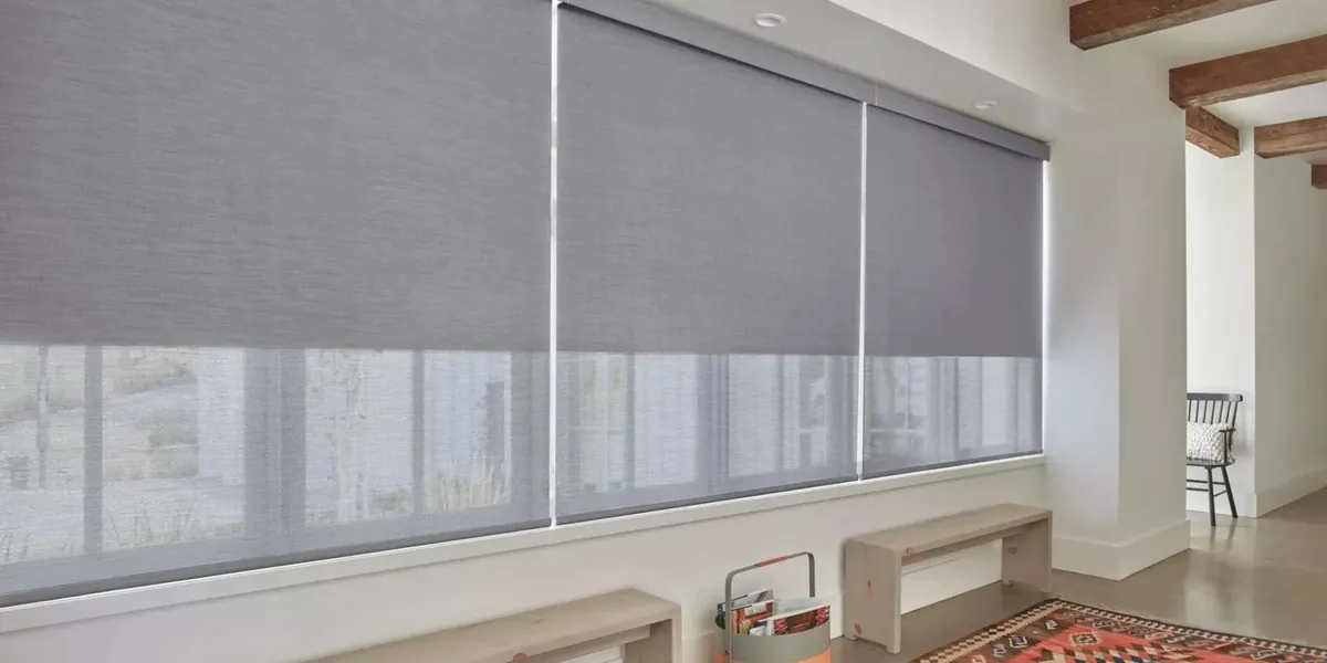 Grey roller shades lowered halfway on a long run of windows in a white hallway with benches and a patterned rug