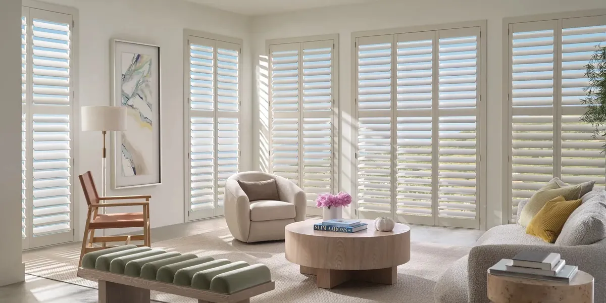 Off-white plantation shutters lining a wall of windows in a sunlit living room with a round wood coffee table
