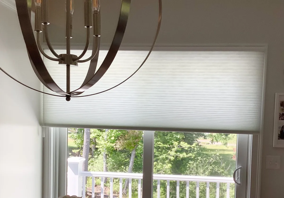 White cellular shade on a sliding glass door to a deck with white railings, trees outside and a round metal chandelier in front