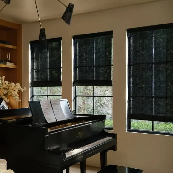 Black woven shades on three tall windows in a music room, with a black grand piano and shelves of books