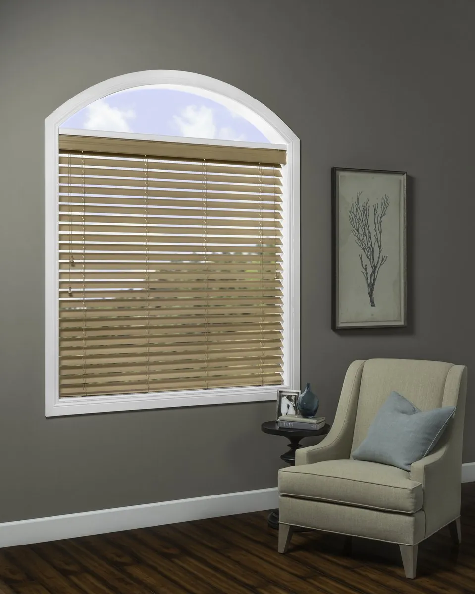 Natural wood blinds lowered on an arch-topped window, the arch left open to the sky, above a grey armchair