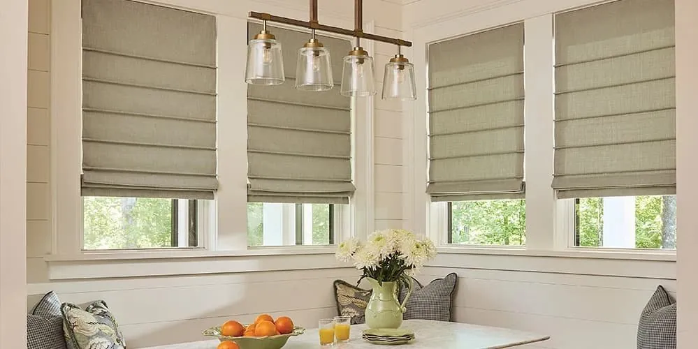 Sage green flat roman shades on breakfast nook windows above a banquette, with pendant lights and a bowl of oranges