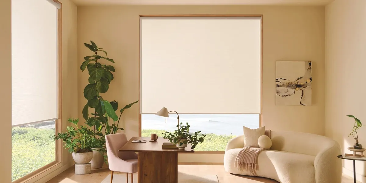 Cream roller shades partly raised on large windows in a home office with a wood desk, curved sofa and ocean view